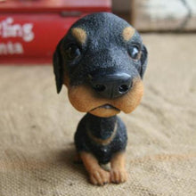Load image into Gallery viewer, A small black and tan rottweiler dog figurine or bobblehead-style toy posed sitting on a beige fabric surface, with a blurred red object in the background.