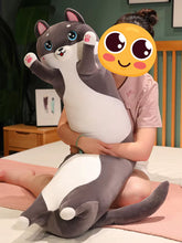 Load image into Gallery viewer, A person sitting on a bed holding a large gray and white husky-shaped plush pillow. The person's face is covered by a yellow cartoon emoji sticker, and the background shows a bedroom setting with a wooden headboard and nightstand.