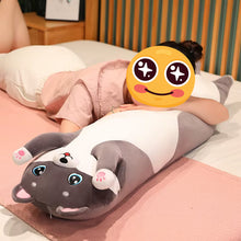 Load image into Gallery viewer, A person lying on a bed with their face obscured by a yellow emoji sticker, resting on a long, plush, husky -shaped body pillow. The pillow features a white torso, gray paws and head, and pink paw pads.