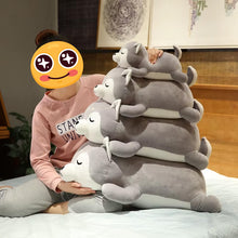 Load image into Gallery viewer, A person sitting on a bed in a bedroom holds a large stack of four plush stuffed animals shaped like gray and white husky dogs. The person's face is covered by a yellow emoji-style sticker.