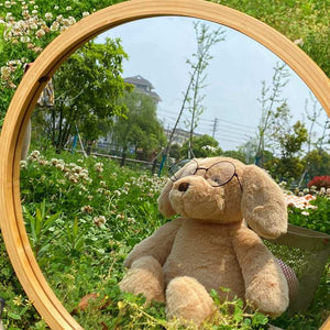 this image shows the cutest plush golden retriever soft toy with an adorable spectacles on , sitting in the  middle of a garden in front of a wooden circular mirror.